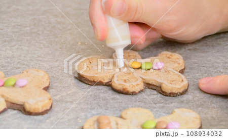 woman's hands decorating a gingerbread man-shaped Christmas cake with white icing and colored caramels, close-up 108934032