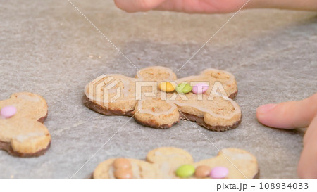 woman's hands decorating a gingerbread man-shaped Christmas cake with white icing and colored caramels, close-up 108934033