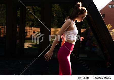 Side view of redhead woman rotating shoulders doing warm up exercise during fitness training, standing on background of house on sunny summer morning. Female training in open air warms joints. 108934683