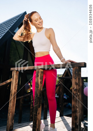 Vertical portrait of charming athletic female in sportswear standing leaning on handrail of country house after training on sunny summer day on background of sky. Sportswoman posing resting outdoors. Vertical portrait of charming athletic female in sportswear standing leaning on handrail of country house after training on sunny summer day on background of sky. Sportswoman posing resting outdoors. 108934686