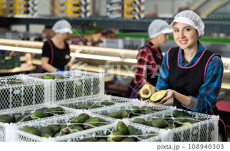 Positive woman with avocado slices in hands at food factory warehouse 108940303