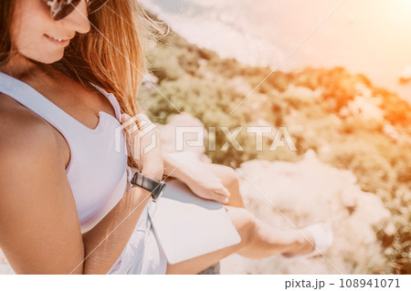 Digital nomad, woman in the hat, a business woman with a laptop sits on the rocks by the sea during sunset, makes a business transaction online from a distance. Freelance, remote work on vacation. 108941071