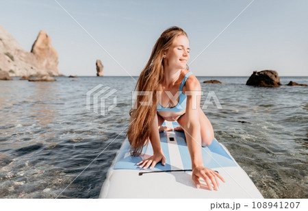 Woman sea sup. Close up portrait of happy young caucasian woman with long hair looking at camera and smiling. Cute woman portrait in a blue bikini posing on sup board in the sea 108941207
