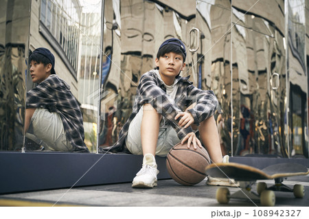 teenage asian kid taking a break sitting on ground with basketball and skateboard teenage asian kid taking a break sitting on ground with basketball and skateboard 108942327
