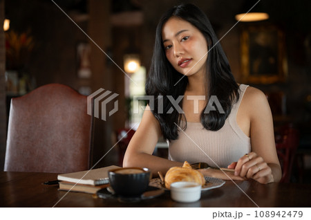 Elegant young Asian woman eating croissant fro breakfast with hot latte coffee in a dark luxury living room. 108942479
