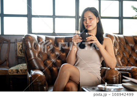 Elegant and classy woman drinking hot latte while sitting on vintage sofa in her living room. 108942499