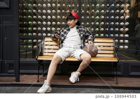 fifteen-year-old teenage asian boy sitting on bench looking at cellphone 108944503