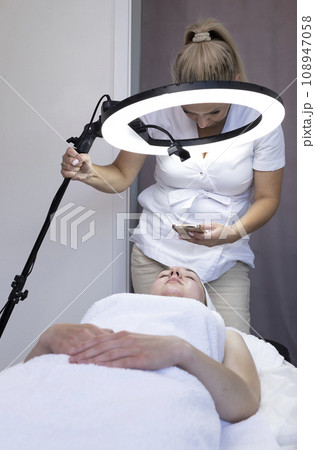 Caucasian Woman Cosmetologist Or Beautician Takes Pictures On Phone Of Cosmetology Procedure Result On Young Beautiful Female Patient In Medical Office, Led Selfie Ring Light on Tripod. Vertical. Caucasian Woman Cosmetologist Or Beautician Takes Pictures On Phone Of Cosmetology Procedure Result On Young Beautiful Female Patient In Medical Office, Led Selfie Ring Light on Tripod. Vertical. 108947058