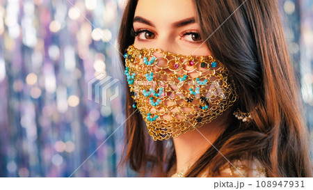 Pandemic fashion. DIY accessory. Closeup of mysterious woman with night makeup in matching glamour gold chain face mask on blur blue sparkling tinsel background. 108947931