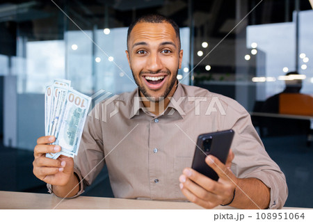 Excited man sitting in modern office holding smartphone and wad of dollars in hands, happy with winning, smiling and looking at camera. 108951064