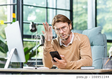 Angry businessman holding phone, shouting and waving hand, sitting at desk in office, upset about bad news, angry at non-working phone. Angry businessman holding phone, shouting and waving hand, sitting at desk in office, upset about bad news, angry at non-working phone. 108951144