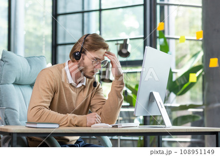 Tired worker in headset holding hand to head, sitting at table, suffering from headache, tired from work wants to sleep. 108951149