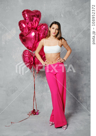 Cheerful young woman in wide bright pink trousers and white top posing next to balloons 108952372