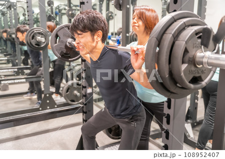 バーベルをつらそうに持ち上げる男性と補助に入るトレーナー バーベルをつらそうに持ち上げる男性と補助に入るトレーナー 108952407