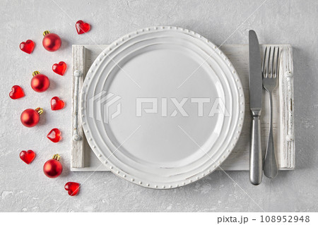 Empty white plate with cutlery on a white wooden tray on a gray concrete table surrounded by red Christmas balls and hearts. Template for presenting food on the menu 108952948