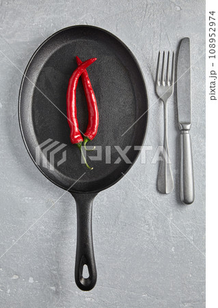 Two hot peppers on a black cast iron frying pan and cutlery on the background of a stone table. Top view with copy space Two hot peppers on a black cast iron frying pan and cutlery on the background of a stone table. Top view with copy space 108952974