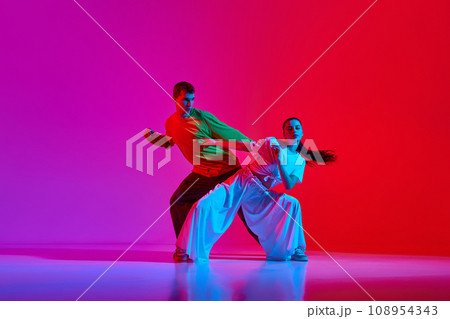 Creative flow. Young man and woman in motion, dancing hip hop against pink red background in neon light Creative flow. Young man and woman in motion, dancing hip hop against pink red background in neon light 108954343