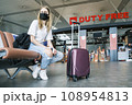 Young teenage girl is sitting at the airport waiting for her flight next to her suitcase. Girl wears a mask Young teenage girl is sitting at the airport waiting for her flight next to her suitcase. Girl wears a mask 108954813