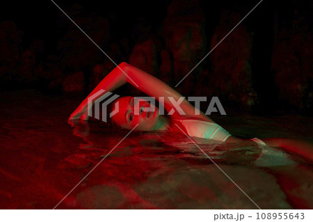 Woman swims in lagoon at night, lit by red light. Female beauty highlighted in natural water pool. Model poses during nocturnal photoshoot. Creative water portrait. Lady in wet swimsuit. Woman swims in lagoon at night, lit by red light. Female beauty highlighted in natural water pool. Model poses during nocturnal photoshoot. Creative water portrait. Lady in wet swimsuit. 108955643