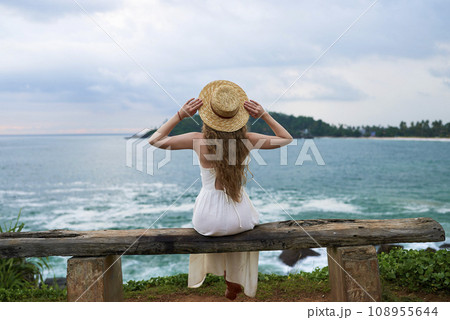 Young woman in white dress sits on rustic bench by the sea, contemplating waves. Back view of female in straw hat enjoys ocean landscape, solitude moment, tropical coast. 108955644