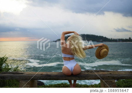 Woman in bikini with a beautiful physique sitting on beach looking at ocean view on sunset in swimwear. Slim figure girl enjoys sea vacation with straw hat in hands. Pretty slender fit blonde female. 108955647