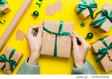 Woman is packing surprise gift for Christmas holidays, sitting at workplace with craft tools, top view. Woman wrapping Christmas gift at colored table 108958886