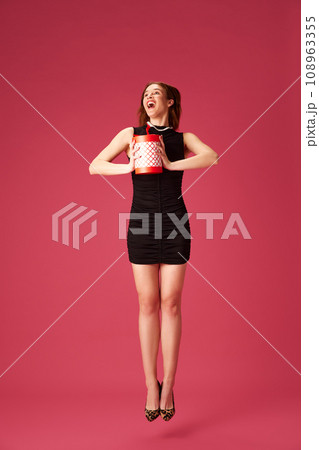 Happy young girl in elegant black dress holding present, celebrating against pink studio background. Birthday Happy young girl in elegant black dress holding present, celebrating against pink studio background. Birthday 108963355