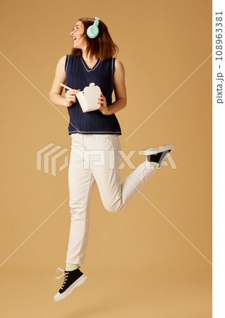 Positive young girl in headphones eating noodles with chopsticks against beige studio background. Dinner to go. 108963381