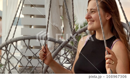 Smiling female tourist enjoying sea breeze while swinging on the beach at sunset. Concept of summertime, vacation and beach holidays. 108967306