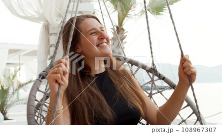 Smiling brunette woman smiling and relaxing in rope nest chair swinging at sea beach bar. Concept of summertime, vacation and beach holidays. Smiling brunette woman smiling and relaxing in rope nest chair swinging at sea beach bar. Concept of summertime, vacation and beach holidays. 108967307