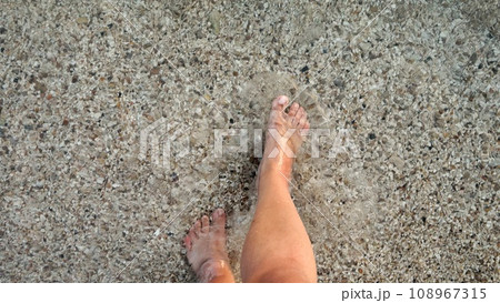 Male feet walking over sandy ocean beach and pebbles through calm clear sea water 108967315