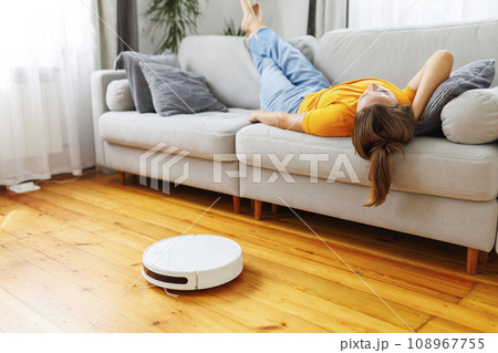 A woman sleeping on sofa while robot vacuum quietly takes care of the cleaning A woman sleeping on sofa while robot vacuum quietly takes care of the cleaning 108967755