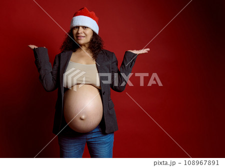 Charismatic pregnant woman, expectant mother in stylish gray blazer and Santa hat, holding copy space on hands palms up 108967891