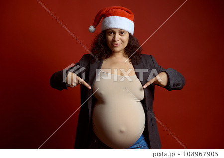 Pregnant woman in Santa hat, pointing fingers at her big belly in pregnancy third trimester, isolated on red background. Pregnant woman in Santa hat, pointing fingers at her big belly in pregnancy third trimester, isolated on red background. 108967905