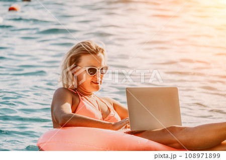Woman works on laptop in sea. Freelancer, young blond woman in sunglases floating on an inflatable big pink donut with a laptop in the sea at sunset. Freelance, travel and holidays concept. Woman works on laptop in sea. Freelancer, young blond woman in sunglases floating on an inflatable big pink donut with a laptop in the sea at sunset. Freelance, travel and holidays concept. 108971899