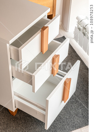 Stylish white office commode with wooden handles and legs with open empty drawers closeup view. Comfortable furniture for home and office furnishing Stylish white office commode with wooden handles and legs with open empty drawers closeup view. Comfortable furniture for home and office furnishing 108978253