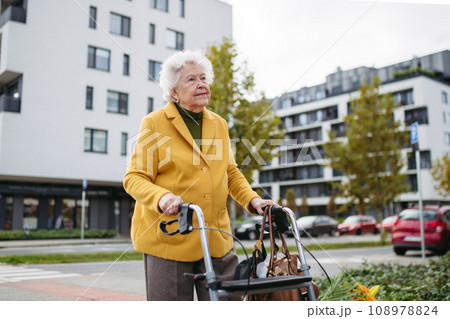 Senior woman with a mobility walker walking on city streets during autumn day, enjoying the beautiful sunny weather. Elderly lady savoring every moment, living life to fullest. 108978824