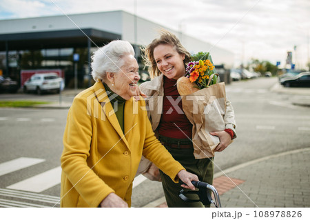 Mature granddaughter carrying grandmother's shopping bag. Senior woman and caregiver going to home with groceries from supermarket, during cold autumn day. 108978826
