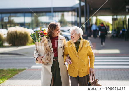 Mature granddaughter carrying groceries out to her grandmother's car. Senior woman shopping at the shopping center, needing help loading groceries into car. 108978831