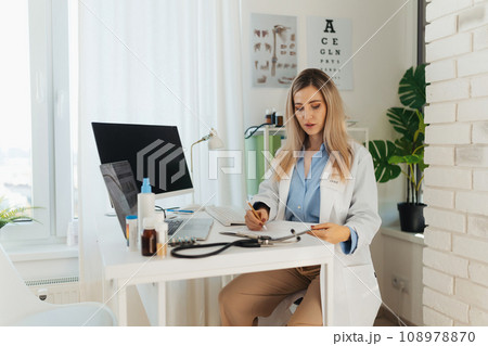 Young beautiful female doctor working on laptop in doctor's office. Physician doing paperwork and administration. 108978870