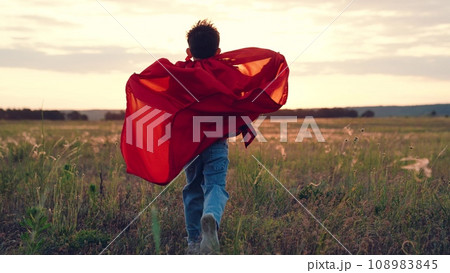 Boy races along field at sunset with raincoat flapping in wind under bright sun Boy races along field at sunset with raincoat flapping in wind under bright sun 108983845