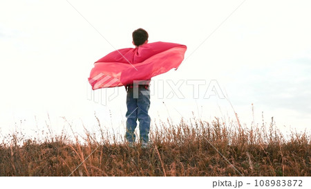 Boy stands on field straightening arms with red raincoat in cloudy weather Boy stands on field straightening arms with red raincoat in cloudy weather 108983872