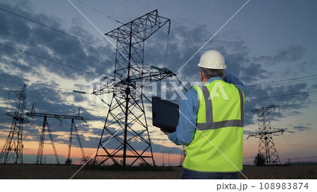 Technician works with network data on laptop walking to power transmission lines Technician works with network data on laptop walking to power transmission lines 108983874