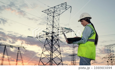 Engineer with helmet holds laptop to control power transmission lines at sunset Engineer with helmet holds laptop to control power transmission lines at sunset 108983894