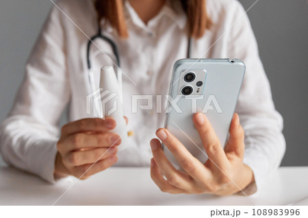 Internet-based medical services. Unrecognizable woman doctor wearing white medical lab coat stethoscope using smart phone for online consultation prescribes spray for patient telling how to take it. 108983996