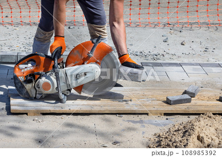 A worker cuts paving slabs with a diamond blade and a gas cutter on a clear summer day. A worker cuts paving slabs with a diamond blade and a gas cutter on a clear summer day. 108985392