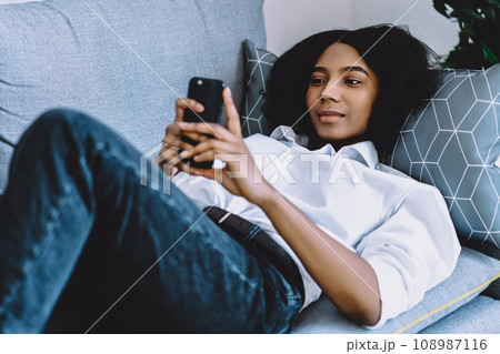 African american hipster girl messaging on mobile phone lying on sofa in flat 108987116
