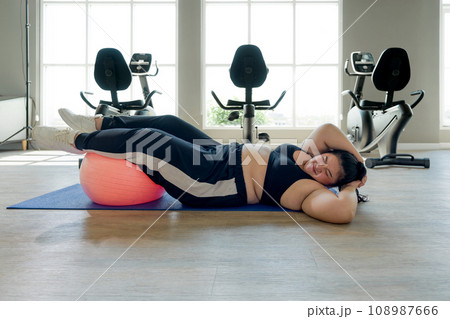 An expressive, slightly overweight woman is exerting herself as she lays on a large exercise ball in a gym. An expressive, slightly overweight woman is exerting herself as she lays on a large exercise ball in a gym. 108987666