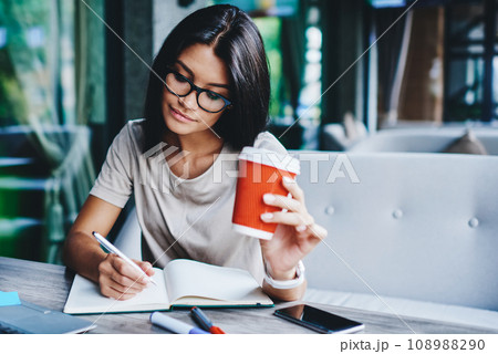 Concentrated female student writing in notebook while learning with cardboard coffee cup in cafe Concentrated female student writing in notebook while learning with cardboard coffee cup in cafe 108988290