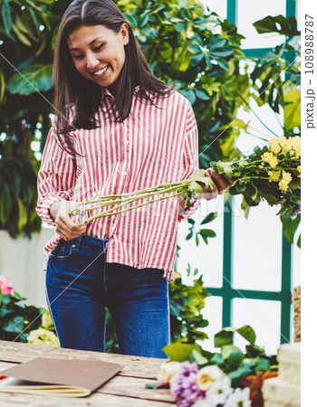 Cheerful asian woman designing floral composition while working in glasshouse near desk Cheerful asian woman designing floral composition while working in glasshouse near desk 108988787